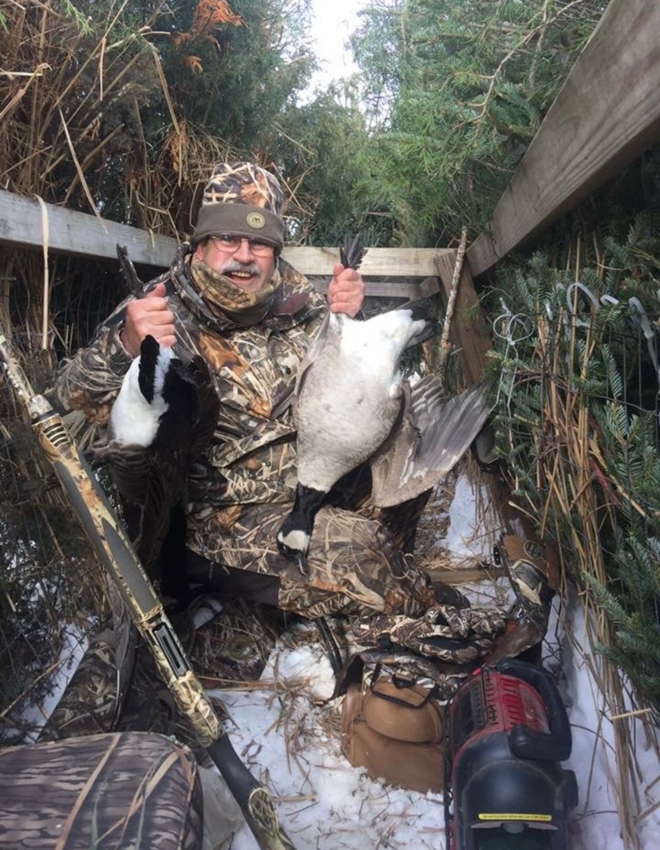 Hunter in a pit blind holding up two Canada geese